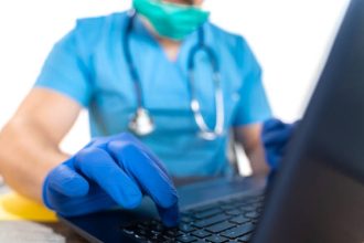 Doctor with stethoscope using his laptop keyboard, white background. The scene is situated in controlled photography studio environment in front of white background. Picture is taken with Sony A7III camera.